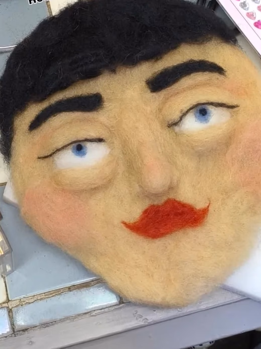 A needle-felted puppet face resting on a workbench beside a makeup palette