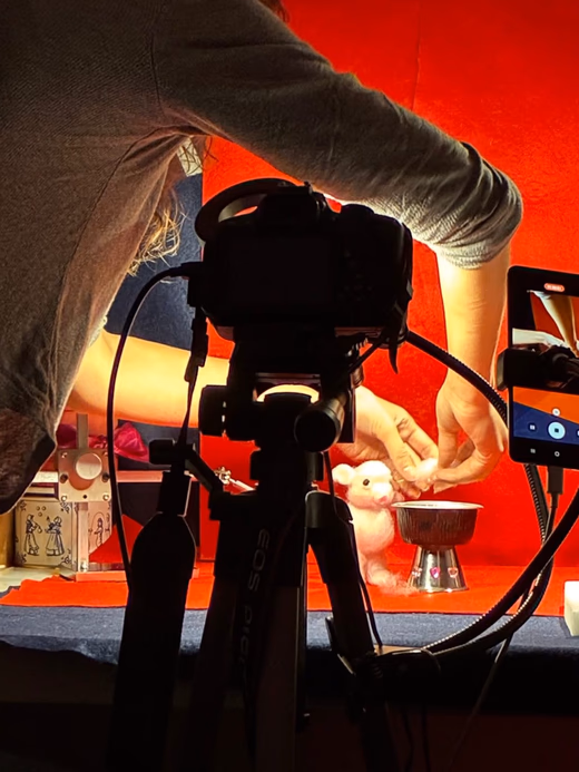 Hands adjusting a needle-felted puppet on a red set, behind a camera mounted on a tripod