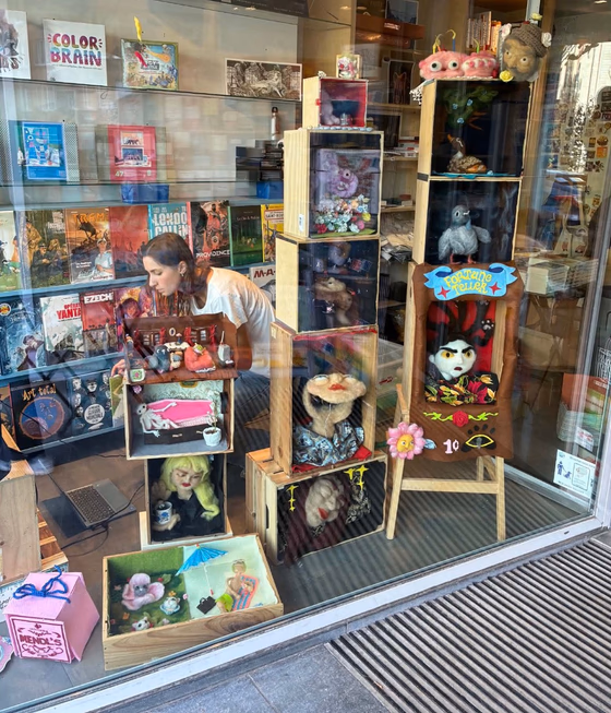 Vitrine de la librairie Bleus d'Encre : marionnettes en laine feutrée dans des caisses en bois empilées, avec un théâtre « Fortune Teller », pendant que Margot termine l'installation
